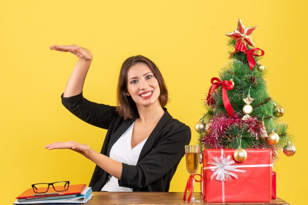 L'humeur de Noël avec de jeunes satisfaits heureux femme d'affaires émotionnelle montrant quelque chose et assis à une table dans le bureau sur jaune