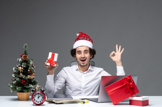 Humeur de Noël avec souriant jeune homme d'affaires avec chapeau de père Noël et tenant son cadeau faisant des gestes de lunettes sur fond sombre