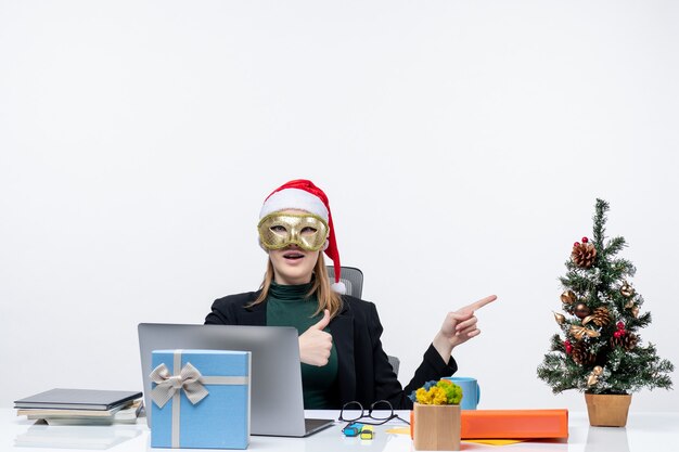 L'humeur de Noël avec surpris jeune femme avec chapeau de père Noël et portant un masque assis à une table pointant quelque chose sur fond blanc