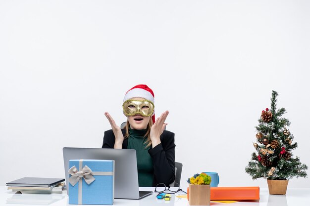 L'humeur de Noël avec surpris jeune femme excitée avec chapeau de père Noël et portant un masque assis à une table sur un fond blanc