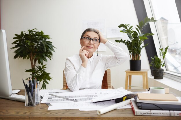 Idées, inspiration et pensée créative. Photo de l'ingénieur femme mûre caucasienne réfléchie à lunettes à la recherche sur le côté avec un sourire mystérieux et pointant le doigt vers le haut comme si ayant une bonne idée