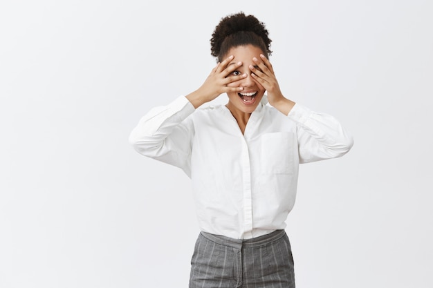 Photo gratuite il est temps de jouer à cache-cache. portrait de joyeuse femme entrepreneur à la peau sombre attrayante et drôle s'amusant, étant enfantine et insouciante tout en jouant à peekaboo, furtivement à travers les doigts et souriant