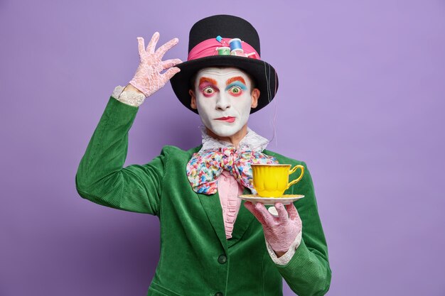 Il est temps de prendre le thé. Un homme aristocratique avec un maquillage lumineux a l'image d'un personnage fictif tient une tasse de boisson porte un grand chapeau s'est demandé des poses d'expression sur un mur violet