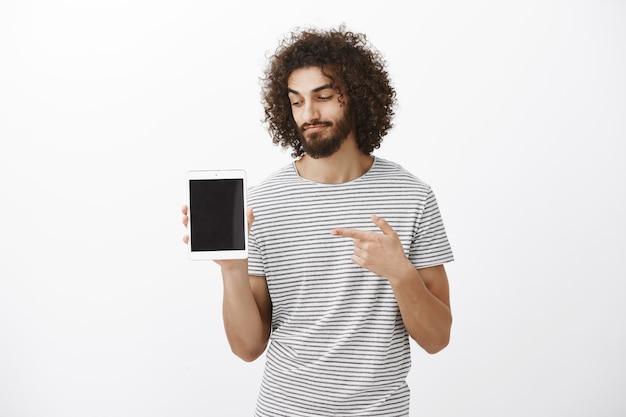 Photo gratuite il sait quels gadgets valent la peine d'être achetés. client masculin satisfait satisfait avec barbe et coupe de cheveux élégante en chemise rayée, pointant et regardant la tablette numérique avec un léger sourire