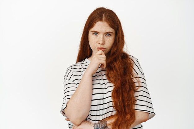 Image d'une adolescente rousse réfléchie plissant les yeux, touchant le menton, considérant qch, pensant, prenant une décision, réfléchissant aux choix, debout sur fond blanc.