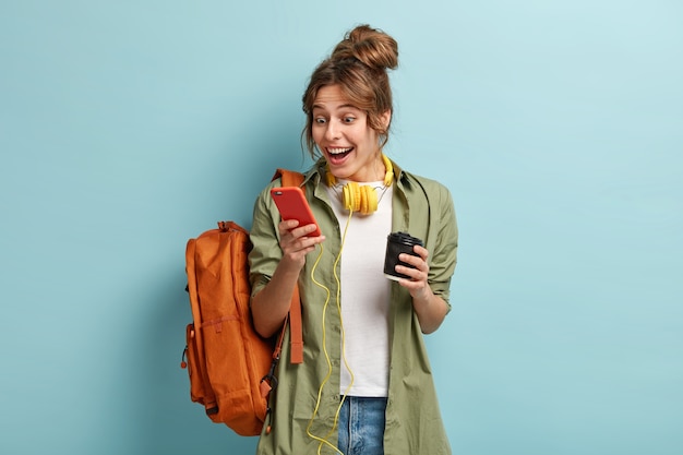 Image d'une adolescente souriante et joyeuse aime la communication avec un ami dans le chat de groupe, regarde des photos drôles en ligne, écoute de la musique hors ligne avec des écouteurs, boit du café aromatique dans une tasse en papier