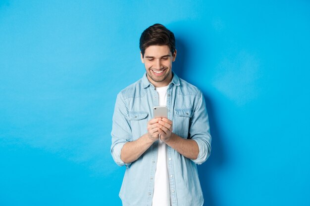 Image d'un beau jeune homme utilisant un téléphone portable, envoyant des SMS au téléphone et l'air heureux, debout sur fond bleu.