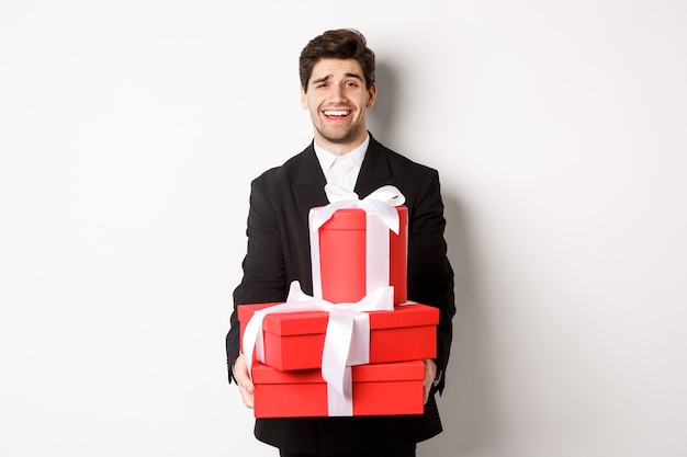 Image de beau mec en costume noir, tenant des cadeaux pour les vacances de Noël, debout sur fond blanc