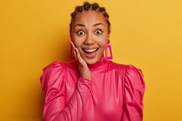 L'image d'un beau modèle à la peau sombre a l'air excité et heureux, touche la joue, porte un chemisier rose élégant, se sent heureux de regarder une nouvelle tenue à la mode en magasin, a un sourire radieux, pose à l'intérieur