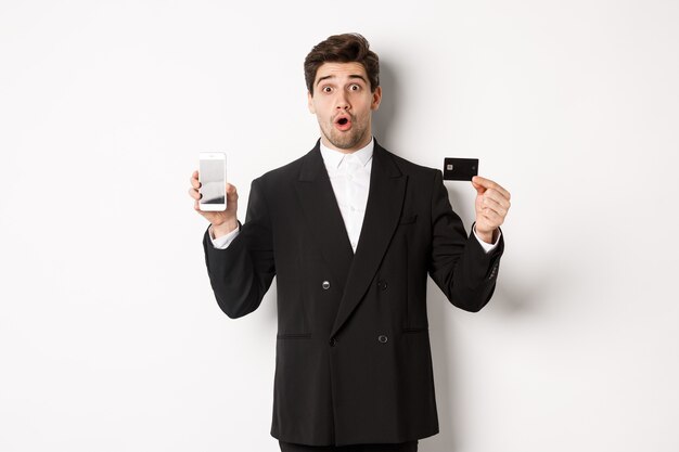 Image d'un bel homme d'affaires en costume noir, l'air étonné et montrant une carte de crédit avec écran de téléphone portable, debout sur fond blanc.