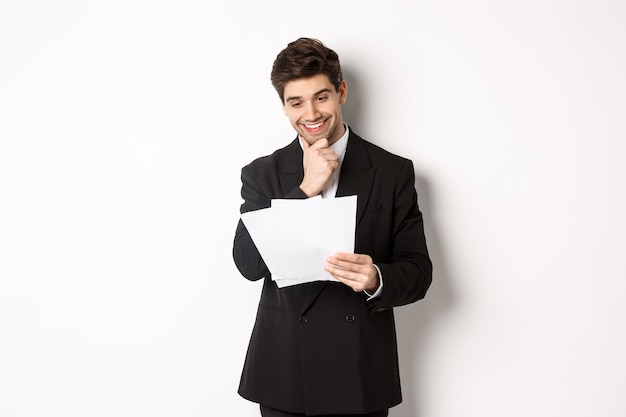 Image d'un bel homme d'affaires en costume noir, l'air satisfait de documents, lisant un rapport et souriant, debout sur fond blanc.
