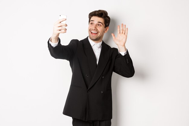 Image d'un bel homme en costume, ayant un appel vidéo et agitant la main à l'appareil photo du smartphone, enregistrant une vidéo, saluant quelqu'un, debout sur fond blanc.