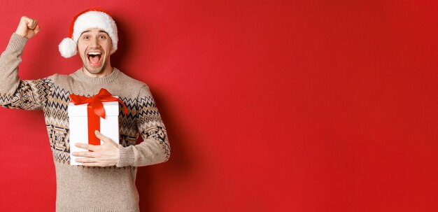 Image d'un bel homme heureux et excité, reglaçant le cadeau de Noël, levant les mains en signe de triomphe et souriant, célébrant le nouvel an, debout sur fond rouge en bonnet de noel