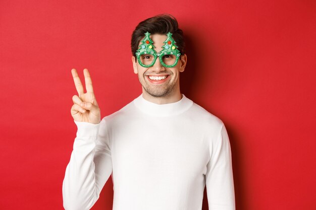 Image d'un bel homme en pull blanc et lunettes de fête, montrant un signe de paix et souriant, souhaitant un joyeux noël, debout sur fond rouge.