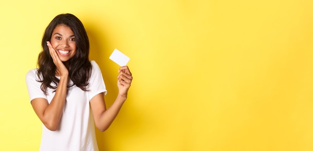 Photo gratuite image d'une belle femme afro-américaine souriante heureuse montrant une carte de crédit debout sur un backgro jaune