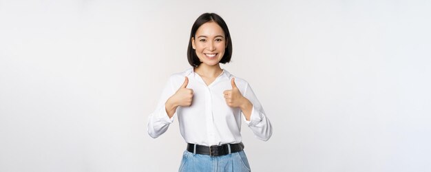 Image d'une belle femme asiatique adulte montrant les pouces vers le haut portant des vêtements universitaires officiels de bureau recommandant une entreprise debout sur fond blanc