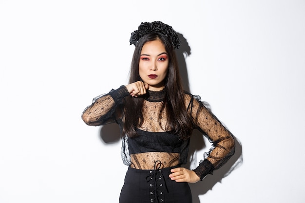 Image de la belle femme asiatique gothique en robe de dentelle noire, menacer quelqu'un, faire un geste de gorge tranchant et regardant sérieusement la caméra, célébrant l'halloween, debout sur fond blanc.