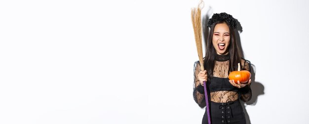 Image d'une belle femme asiatique habillée en sorcière pour la fête d'halloween tenant un balai et une citrouille st