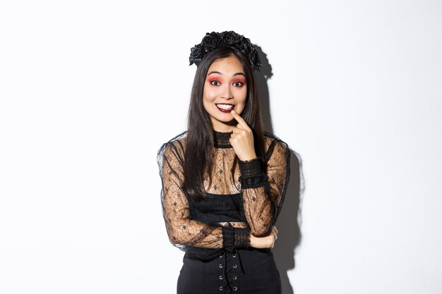 Image d'une belle femme asiatique réfléchie en costume de fête d'halloween à la recherche amusée et intriguée, debout sur fond blanc portant une robe gothique et une couronne.