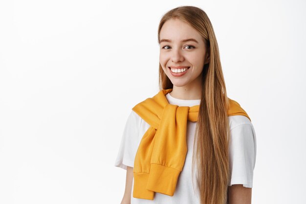 Image d'une belle femme blonde souriante avec de longs cheveux naturels droits, debout à moitié tourné, regardant la caméra avec un visage heureux, fond blanc