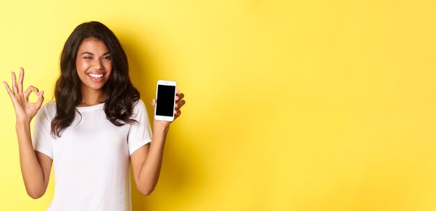 Photo gratuite image d'une belle fille afro-américaine clignant des yeux et souriante montrant l'écran du smartphone et le signe ok