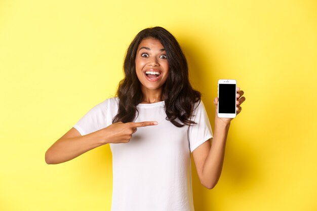 Image d'une belle fille afro-américaine souriante et semblant excitée en pointant sur un smartphone