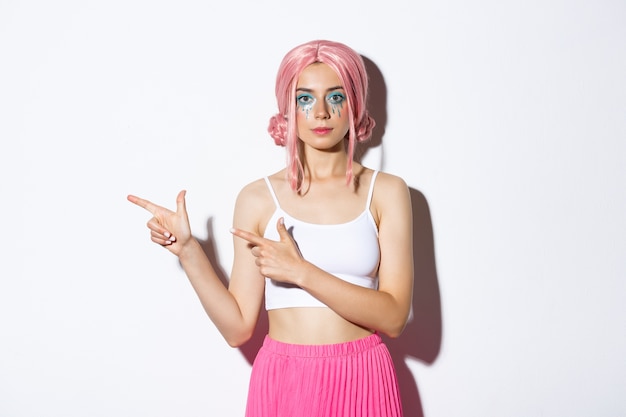 Image de belle fille caucasienne en perruque de fête rose et maquillage lumineux pointant les doigts à gauche sur votre logo, montrant une bannière sur les vacances ou halloween, debout.