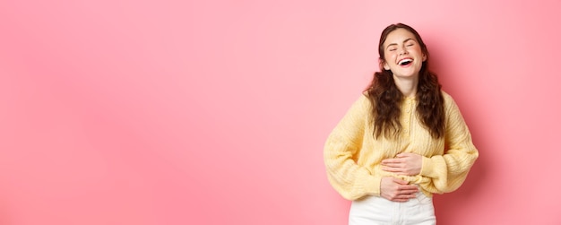 Photo gratuite image d'une belle fille insouciante riant à haute voix en touchant son ventre tout en riant d'une comédie drôle blague hilarante debout sur fond rose