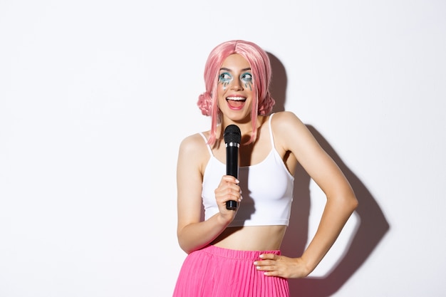 Image de la belle fille souriante en perruque rose, chantant la chanson dans le microphone, portant le costume d'halloween pour la fête, debout.
