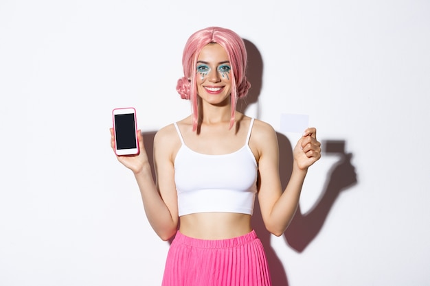 Image de belle fille souriante en perruque rose, avec un maquillage lumineux, montrant la carte de crédit et l'écran du téléphone portable, debout.