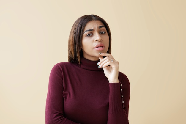 Image de la belle jeune femme afro-américaine douteuse levant un sourcil et touchant le menton, se sentant indécise ou méfiante, regardant avec les yeux pleins de doutes, de désarroi et de suspicion