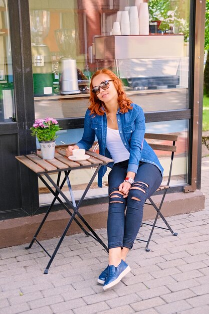 Image complète du corps d'une femme rousse boit du café à la table dans un café dans une rue.