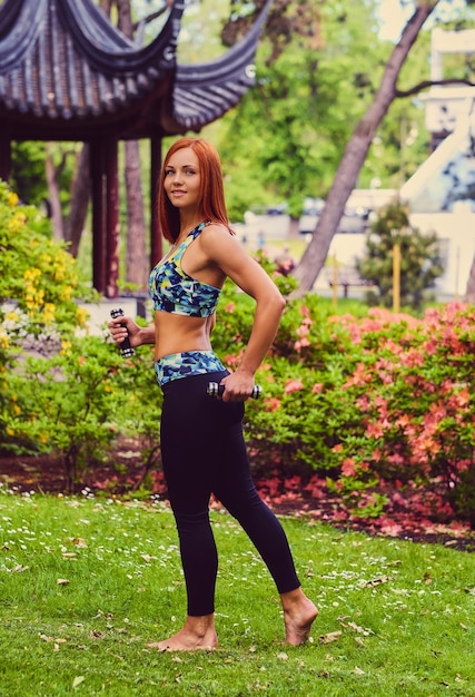 Image complète du corps d'une femme rousse sportive avec des haltères dans un parc extérieur.