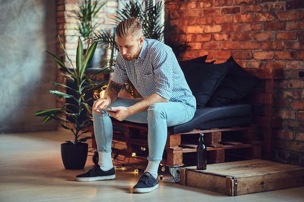 L'image complète du corps d'un homme élégant blond vêtu d'une chemise polaire et d'un jean utilisant une tablette PC dans un salon.