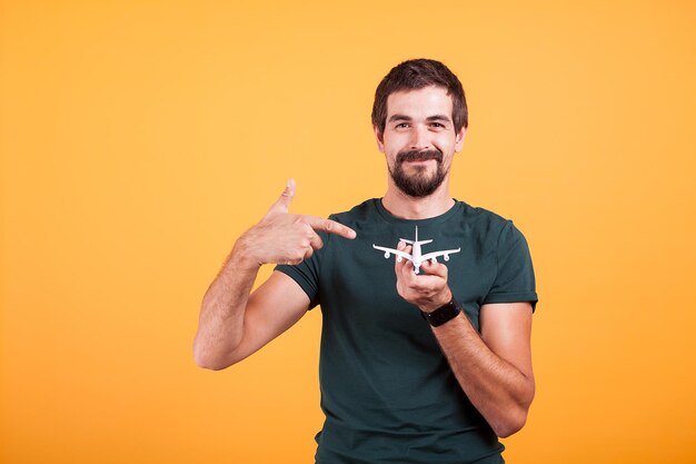 Image de concept de voyage de portrait d'homme pointant vers un avion jouet qu'il tient dans ses mains isolé sur fond orange en studio. Image de vacances, de voyage et de voyage d'été