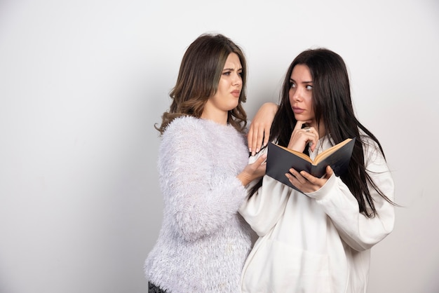 Image de deux meilleurs amis debout ensemble et posant avec un livre sur un mur blanc.