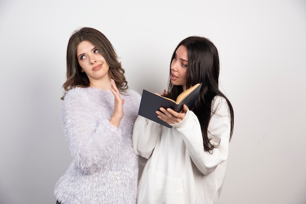 Image de deux meilleurs amis debout ensemble et posant avec un livre sur un mur blanc.