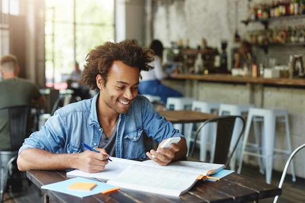 Image d'élégant étudiant africain avec boucle d'oreille portant une chemise en jean assis à une table en bois à faire ses devoirs holding smartphone étant heureux de recevoir un message de son ami en tapant quelque chose
