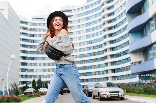 Image extérieure d'une fille joyeuse en chapeau de laine noire et pull d'automne gris sautant et appréciant la promenade dans la ville urbaine moderne.