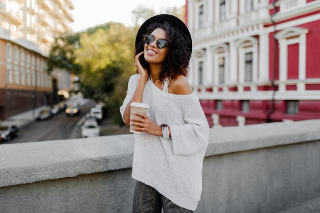 Image extérieure de style de vie aux tons doux de femme noire heureuse qui marche dans la ville de printemps avec une tasse de cappuccino ou de thé chaud. Tenue de hipster. Pull oversize blanc, chapeau noir, accessoires stylés.