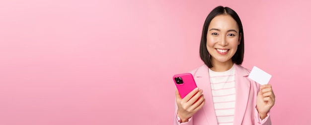 Image D'une Femme D'affaires Asiatique Heureuse Et Souriante Montrant Une Carte De Crédit Payant En Ligne Sur Une Commande D'application Pour Smartphone Avec Un Téléphone Portable Debout Sur Fond Rose