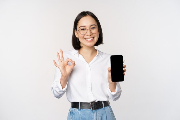 Image d'une femme d'affaires asiatique montrant l'interface de l'application d'écran de smartphone et le signe ok recommandant l'application sur un téléphone mobile debout sur fond blanc