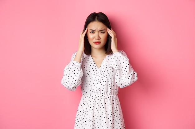 Image d'une femme asiatique contrariée ayant des maux de tête, se sentant mal ou ayant des vertiges, fermer les yeux et se masser la tête, souffrant de migraine, debout sur fond rose.