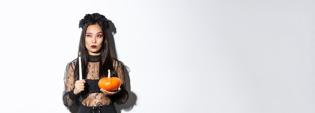 Photo gratuite image d'une femme asiatique en costume de sorcière méchante regardant à gauche sérieuse tenant une bougie allumée et une citrouille cel