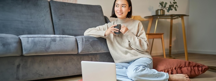 Photo gratuit image d'une femme asiatique souriante buvant du thé chaud tenant une tasse et assise près de l'ordinateur portable sur le sol se reposant