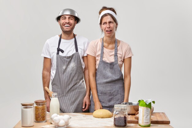 L'image de la femme au foyer surchargée a une expression malheureuse, le mari se tient à côté, aide à faire une tarte au four, prépare de la pâte, prépare un dessert, se tient dans la cuisine, entouré d'ingrédients ou de produits