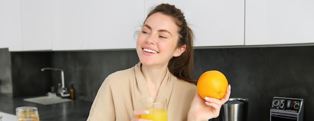 Photo gratuite image d'une femme en bonne santé en peignoir buvant du jus frais montrant des fruits orange posant dans