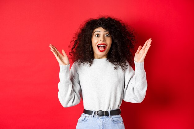 Image d'une femme bouclée heureuse et surprise en maquillage et en sweat-shirt, levant les mains et se réjouissant de la bonne nouvelle, debout sur fond rouge.