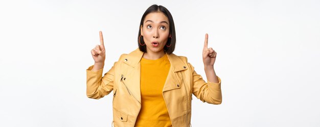 Image d'une femme brune asiatique souriante pointant les doigts vers le haut montrant une publicité avec un visage heureux posant sur fond blanc