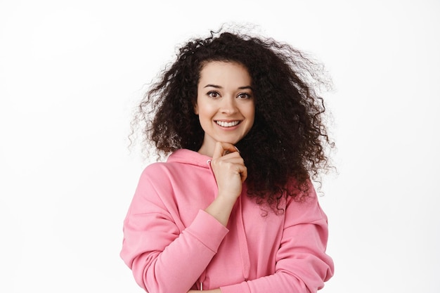 Image d'une femme brune confiante touchant le menton, souriant et regardant la caméra déterminée, debout en sweat à capuche sur fond blanc, a une idée intéressante.